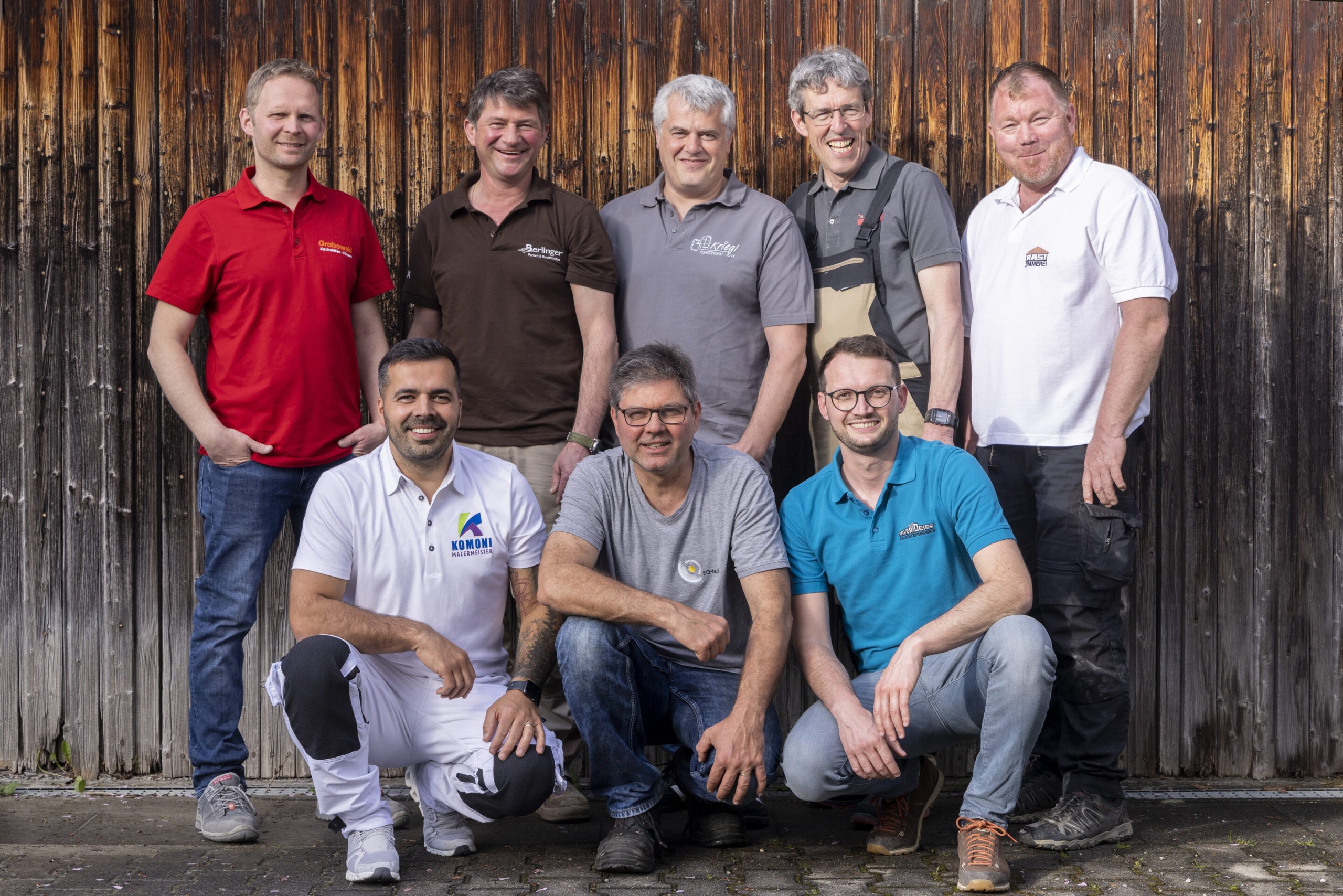 Acht Handwerker in Firmenpoloshirts posieren gemeinsam vor einer verwitterten Holzwand, drei davon in der Hocke