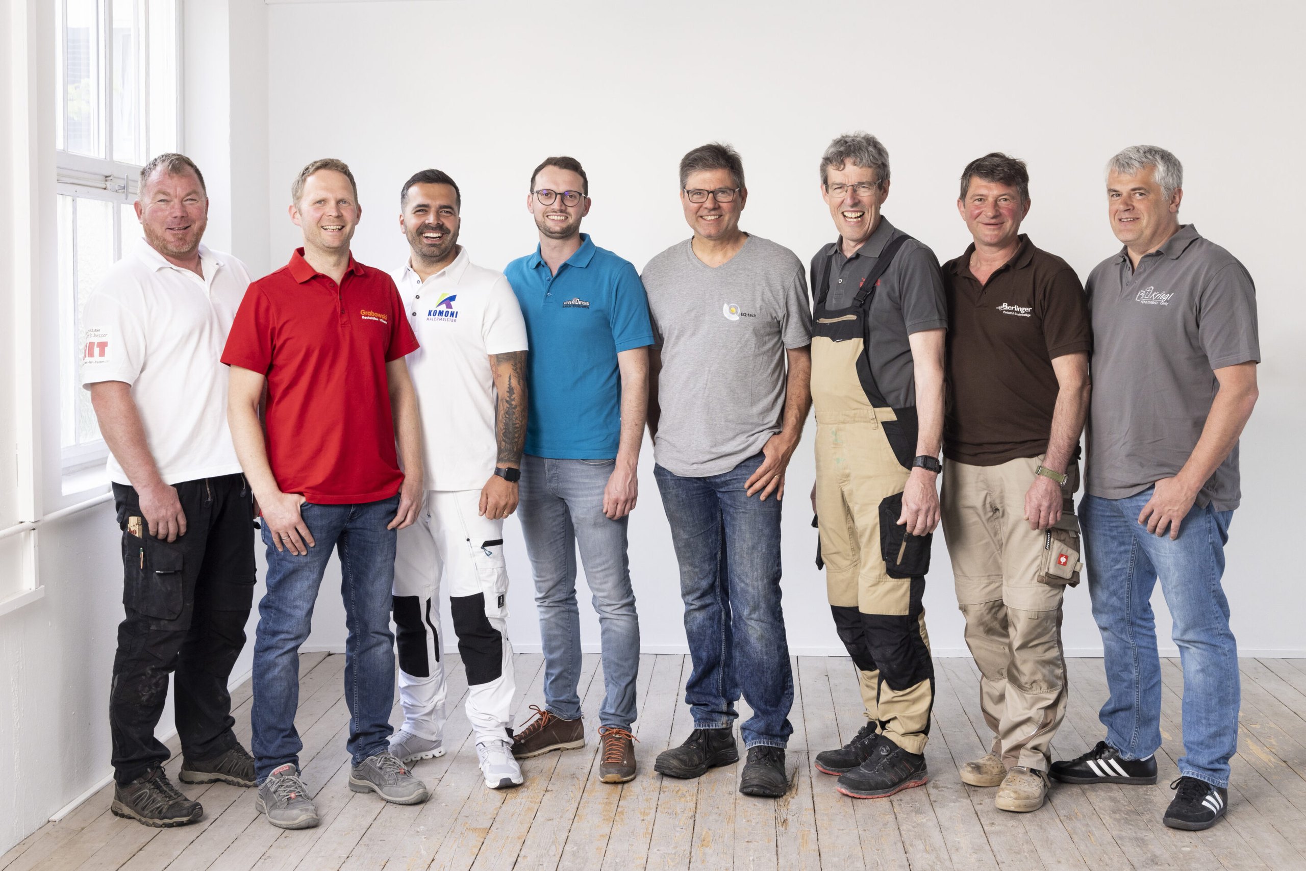 Acht lächelnde Handwerker in Firmenpoloshirts und Arbeitshosen stehen nebeneinander in einem hellen Raum mit Holzdielenboden