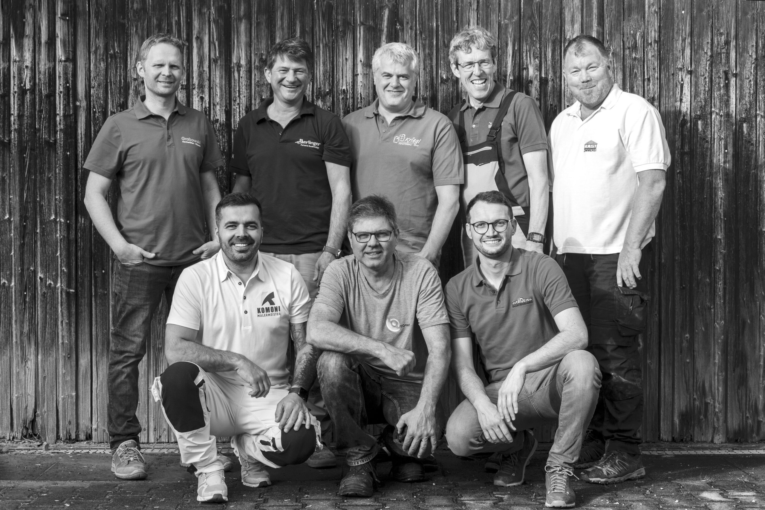 Schwarz-Weiß-Gruppenfoto von acht Handwerkern in Firmenpoloshirts vor einer Holzwand, drei davon in der Hocke