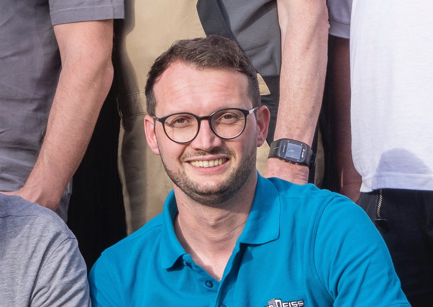 Lächelnder Mann mit runder Brille im türkisblauen Poloshirt, hinter ihm stehende Kollegen in Arbeitskleidung