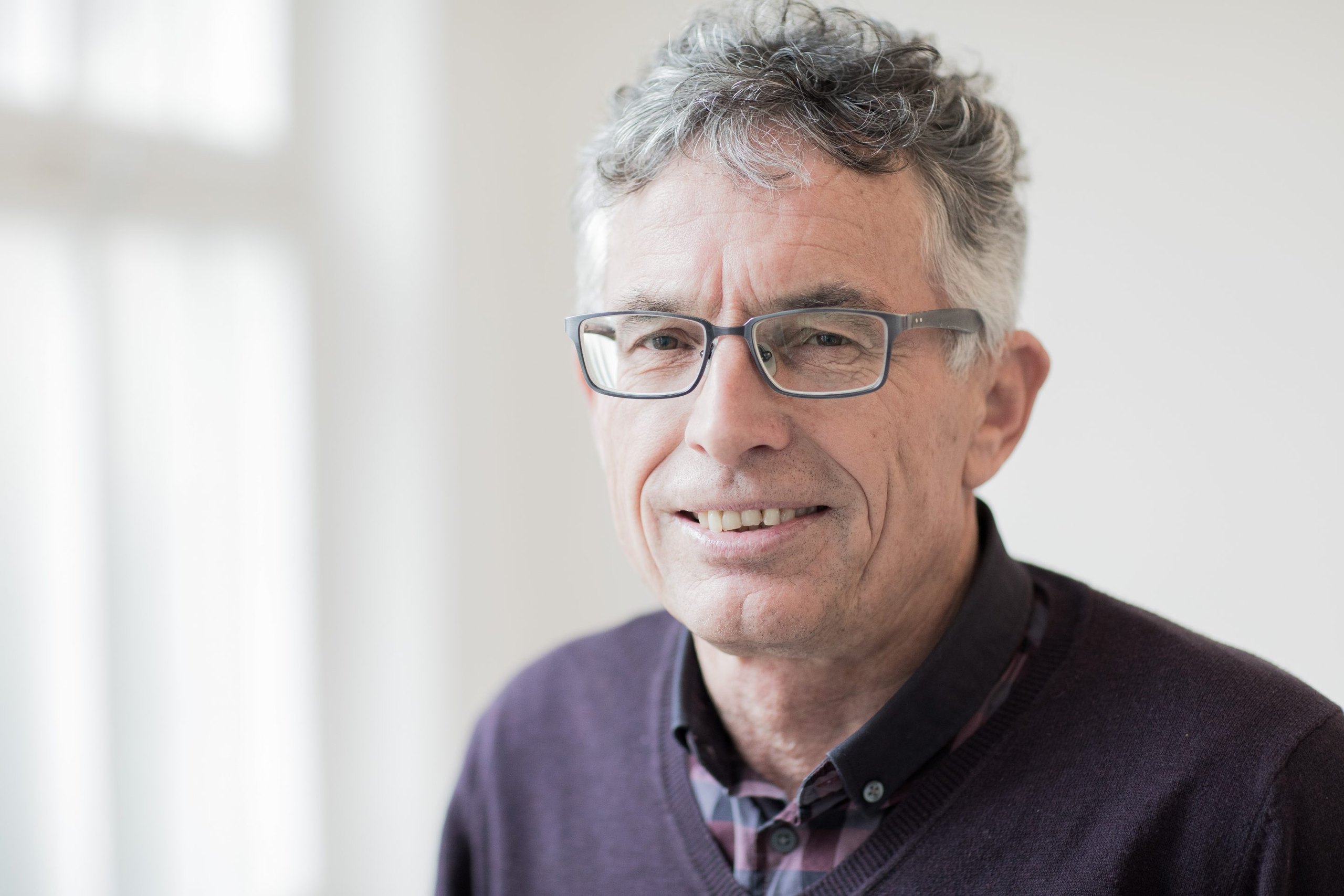 Porträt eines Mannes mit grauen Locken, Brille, violettem Pullover und kariertem Hemd vor hellem Fenster