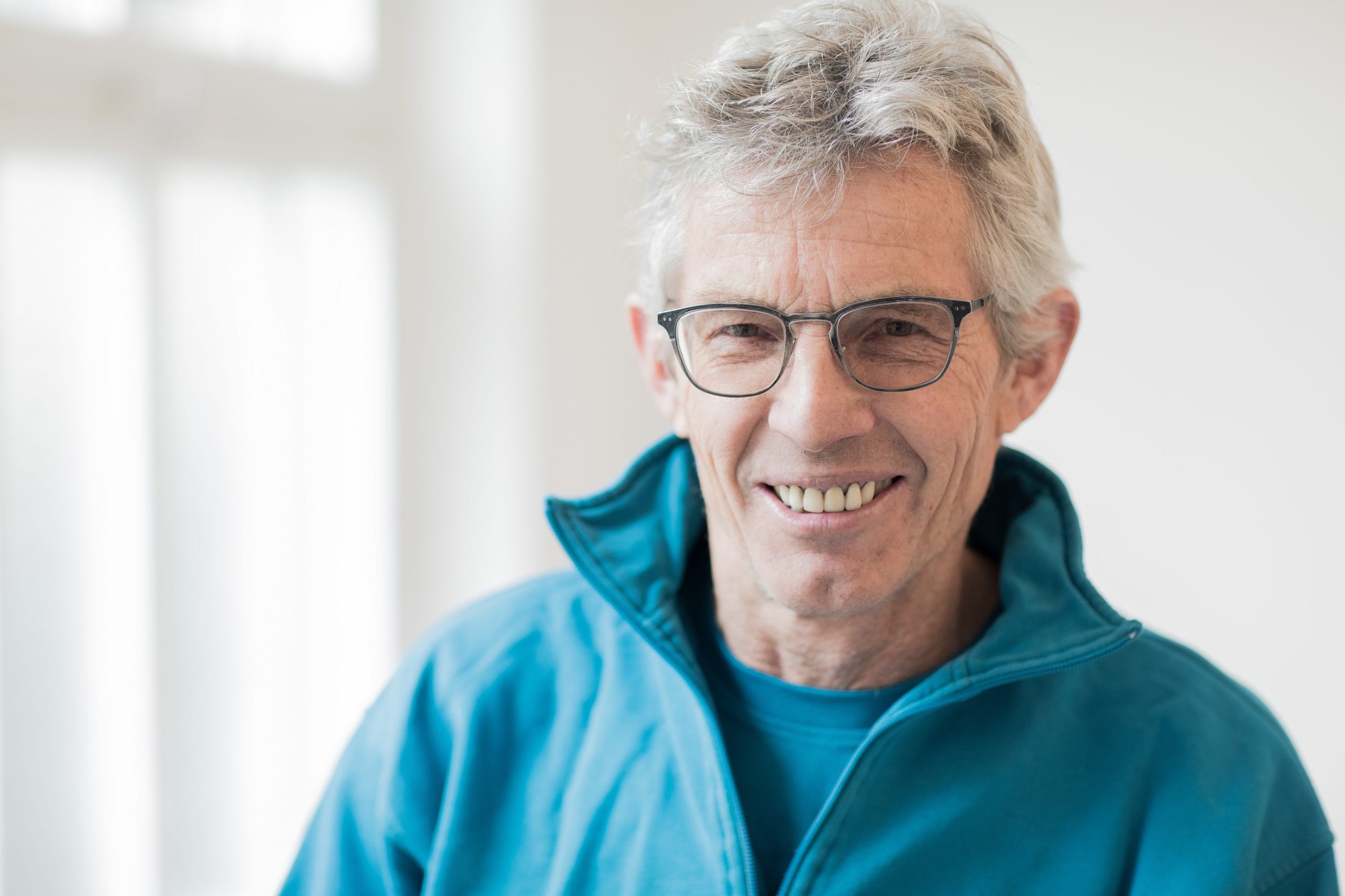 Porträt eines lächelnden Mannes mit grauem Haar und Brille in türkisfarbener Jacke vor hellem Fenster